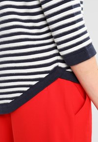 Gestreiftes Oberteil in Marineblau und Weiß, mit abgerundetem Saum und umgeschlagenen Ärmeln, kombiniert mit roten Hosen. Der Stoff wirkt weich und leicht.