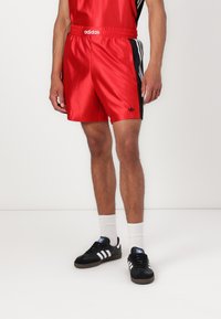 Red satin sports shorts with black side stripes, elastic waistband, and Adidas logo. Paired with black sneakers and white socks.