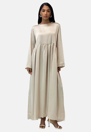 Vestido largo beige de manga larga, hecho de una tela suave, con una cintura fruncida y una falda fluida. Se lleva con tacones blancos y grandes pendientes.