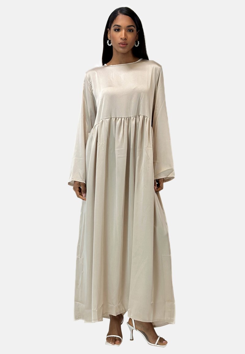 Vestido largo beige de manga larga, hecho de una tela suave, con una cintura fruncida y una falda fluida. Se lleva con tacones blancos y grandes pendientes.