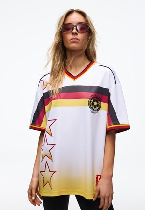 Frau trägt ein weißes Fußballtrikot mit roten, gelben und schwarzen Streifen, Sternmotiven, deutschem Text und rot getönten Sonnenbrillen.
