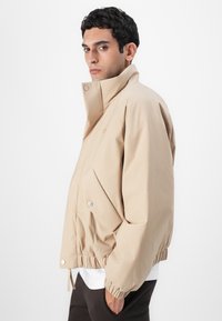 Beige puffer jakk, millel on kõrge krae, elastne alumine serv ja nupuga kinnitused; omab sujuvat tekstuuri ja nurga all külgõmblusi.