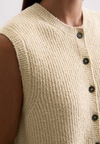 Beige ärmellose Strickjacke aus strukturiertem Stoff mit vertikalen Rippungen, runder Ausschnitt und schwarzen Knöpfen vorne.