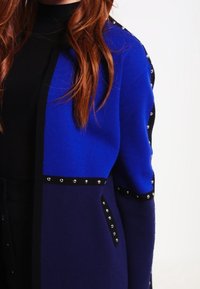 Blå och svart stickad cardigan med silverbeslag längs ärmarna, som har en strukturerad design och kontrasterande färgblock.