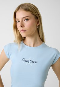 Lichtblauw aansluitend T-shirt met korte mouwen, voorzien van een donkerblauw geborduurd "Guess Jeans" logo en sterontwerp, met een gladde stoftextuur.