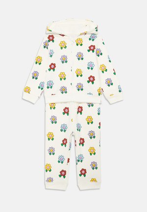 Fato de treino com capuz e fecho éclair em creme, apresentando padrões florais coloridos com rostos sorridentes. Feito de material suave, inclui punhos e cintura elásticos.