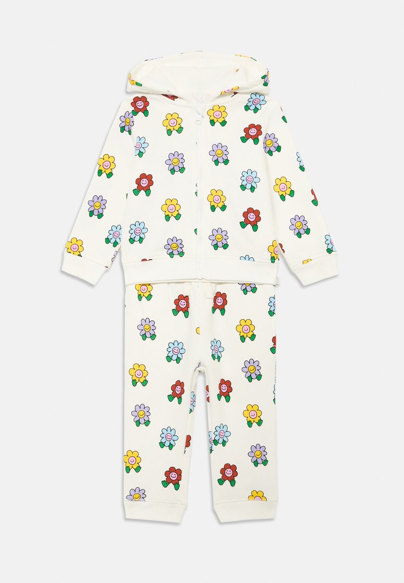 Φόρμα με κουκούλα και φερμουάρ σε κρεμ χρώμα, διακοσμημένη με πολύχρωμα λουλουδάτα μοτίβα και χαμογελαστά πρόσωπα. Κατασκευασμένη από μαλακό υλικό, περιλαμβάνει ελαστικές μανσέτες και μέση.