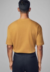 Kortærmet sennepsfarvet t-shirt med en afslappet pasform, glat tekstur og rund halsudskæring, båret med sorte skræddersyede bukser med baglommer.