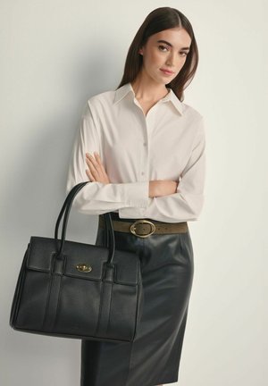 Kobieta z prostymi ciemnymi włosami nosi białą bluzkę, czarną skórzaną spódnicę, trzyma czarną skórzaną torebkę ze złotym zapięciem, stoi na jasnym tle.