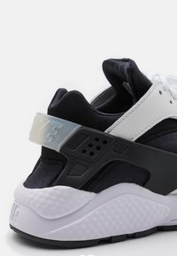 Talons de baskets Nike noir et blanc avec semelle blanche texturée, sangle de maintien en plastique noir et logo blanc sur une sangle semi-transparente.