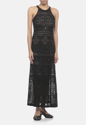 Donna che indossa un abito lungo nero in pizzo senza maniche con motivi geometrici e scarpe nere aperte sulle dita con tacco basso su uno sfondo semplice.