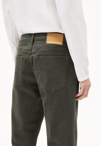 Olijfgroene jeans met achterzakken, gestikte details en een bruine leren patch met het label "ARMEDANGELS" op de tailleband.