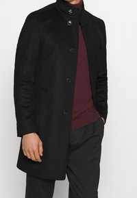 Cappotto di lana nero con colletto alto, dotato di cinque bottoni frontali e tasche laterali. Indossato sopra un maglione bordeaux e pantaloni scuri.