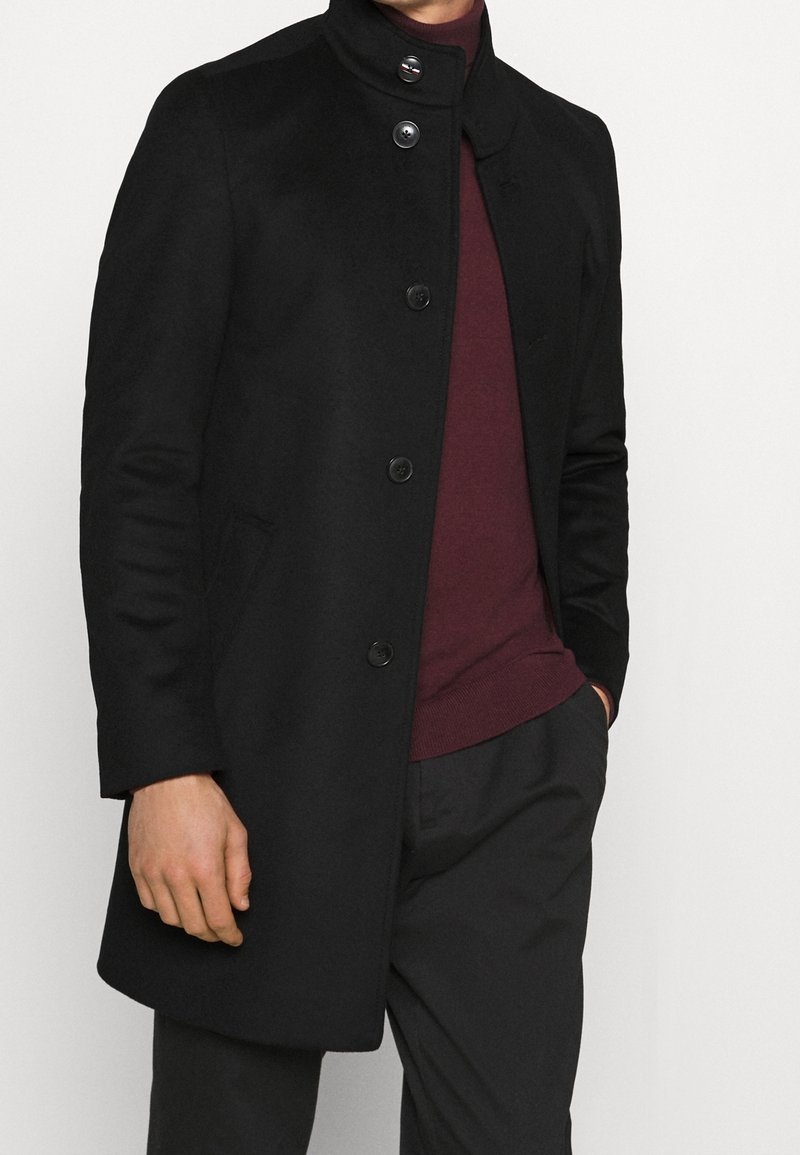 Cappotto di lana nero con colletto alto, dotato di cinque bottoni frontali e tasche laterali. Indossato sopra un maglione bordeaux e pantaloni scuri.