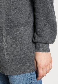 Grau gestrickter Pullover aus Wolle mit gerippten Bündchen, lockerer Passform und einer Fronttasche. Nahaufnahme zeigt die Textur und eine Hand, die auf Denim ruht.