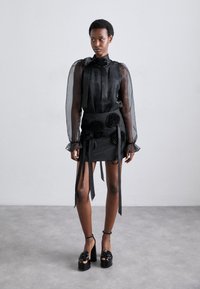 Blouse noire transparente avec un nœud, associée à une mini-jupe noire ajustée ornée d'appliqués de roses et de liens. Portée avec des talons plateformes.
