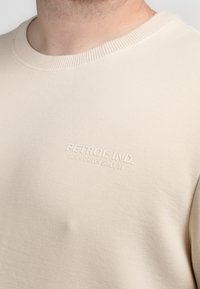 Beige gebreide trui met een geribde ronde hals. Bevat subtiele reliëfbranding aan de voorkant. Gemaakt van katoen of een katoenmix.