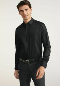 Czarna koszula z długim rękawem, zapinana na guziki, o dopasowanym kroju i subtelnym logo. Noszona z ciemnymi spodniami i czarnym paskiem, eksponując gładką teksturę.