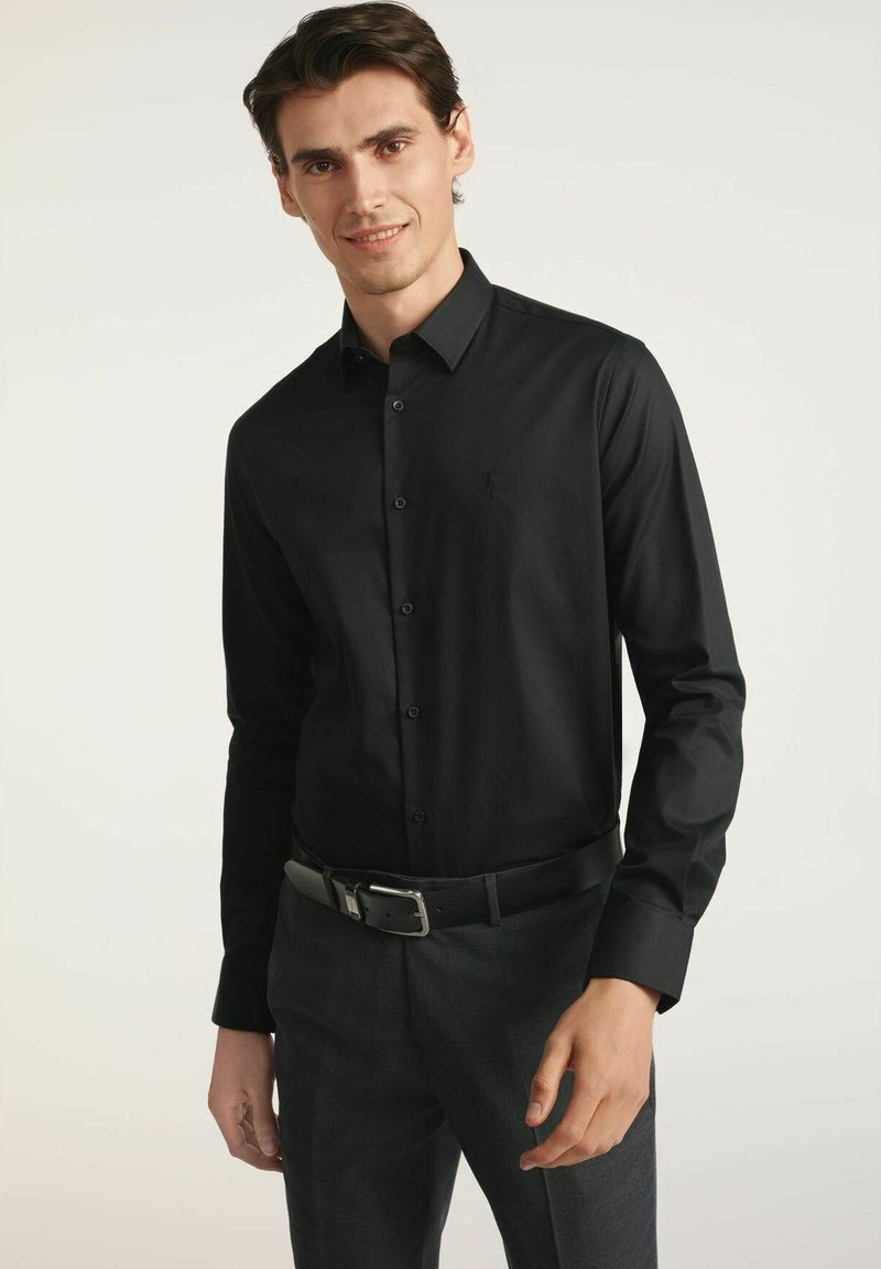 Czarna koszula z długim rękawem, zapinana na guziki, o dopasowanym kroju i subtelnym logo. Noszona z ciemnymi spodniami i czarnym paskiem, eksponując gładką teksturę.