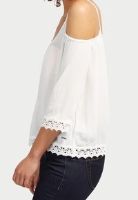 Biała bluzka odkrywająca ramiona z rękawami 3/4, wykonana z teksturowanej tkaniny, ozdobiona koronkową lamówką na dole i przy rękawach. Casualowy fason z cienkimi ramiączkami.
