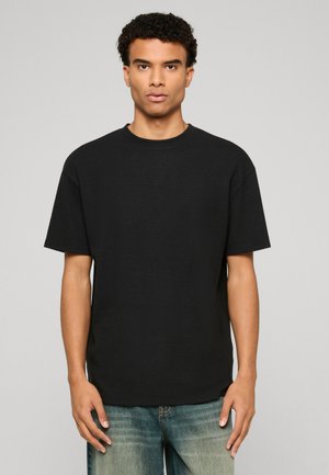 Jeune homme aux cheveux bouclés portant un T-shirt noir uni à manches courtes et un jean bleu délavé, se tenant devant un fond gris.