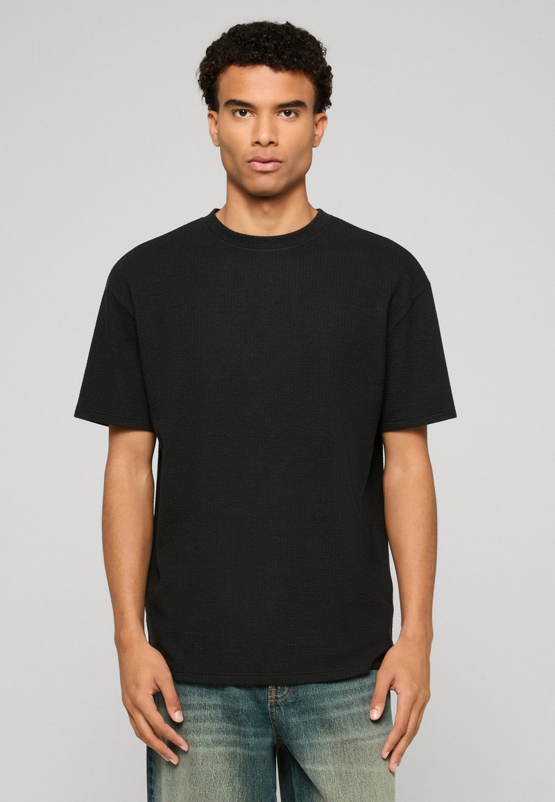 Jeune homme aux cheveux bouclés portant un T-shirt noir uni à manches courtes et un jean bleu délavé, se tenant devant un fond gris.