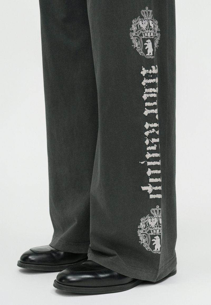 Sackiga mörkgrå byxor med kontrasterande vita grafiska tryck, med en kron- och björndesign längs sidan. Slät textur. Svarta skor syns.