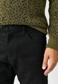 Čierne denimové rifle s prednou kapsou, zabezpečené čiernym gombíkom. Skombinované s chlpatejším zeleným svetrom s čiernym škvrnitým vzorom.