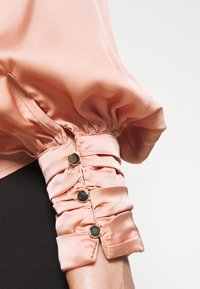 Blusa in satin pesca con maniche a sbuffo e dettagli arricciati, impreziosita da tre bottoni in tono dorato. Texture morbida, forma voluminosa e design con polsini sartoriali.