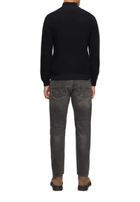 Schwarzer Strickpullover mit hohem Kragen, kombiniert mit grauen Jeans. Auffällige Details sind die gerippten Bündchen und die glatte Textur. Braune Stiefel getragen.