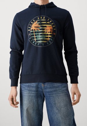 Granatowa bluza z kapturem ze sznurkami i grafiką palmy otoczoną napisem "Relax and Enjoy Jack & Jones 1990", noszona z niebieskimi dżinsami.
