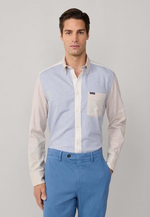Camicia button-up con pannelli azzurri e beige, dotata di taschino sulla sinistra e etichetta con logo, abbinata a pantaloni azzurri.