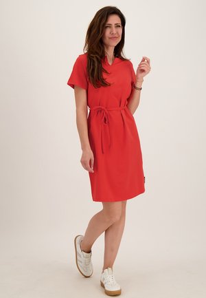 Vrouw die loopt en een knielange rode jurk draagt met korte mouwen en een tailleband, gecombineerd met witte sneakers op een effen achtergrond.