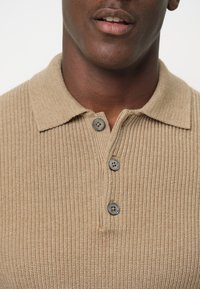Beige ribbed Strickpolohemd mit Kragen, drei dunkelgrauen Knöpfen und einer strukturierten Stoffoberfläche. Minimalistisches Design ohne sichtbare Logos.