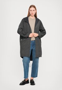 Zwarte patroon buitenjas met een ritssluiting, grote capuchon en twee zijzakken. Gedeeltelijk over een lichtgrijze top en blauwe jeans gedragen.
