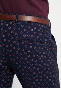 Marineblauwe broek met een diamanten patroon in bordeauxrood, voorzien van een bruine riem. De stof lijkt een textuur te hebben met een zijzak met rits.