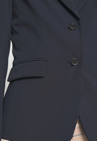 Gros plan sur un blazer bleu marine foncé avec deux boutons noirs et une poche à rabat sur le côté gauche, porté sur un tissu rayé.