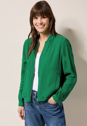 Veste bomber verte en tissu texturé, fermeture éclair, col montant et poche avant. Portée sur un haut blanc et assortie avec un jean bleu.