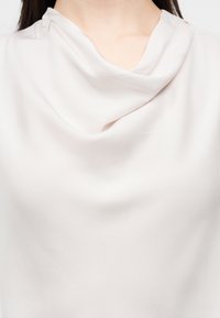 Ženska, oblečena v belo bluzo z mehkim, padajočim ovratnikom v obliki kapuce, ki razkriva vrat in zgornji del prsnega koša na enakomernem ozadju.