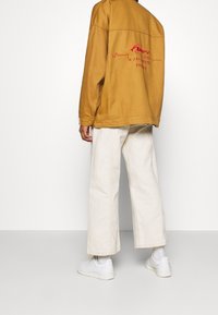 Gul-brun oversized jacka med röd broderad text på ryggen, parat med krämfärgade vida byxor och vita sneakers.