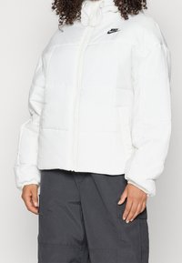 Vit isolerad pufferjacka med hög krage, framdragkedja och svart Nike-logga. Har ett quiltat mönster och elastiska mudd.