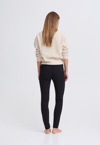 Beige pulóver felgöngyölt ujjakkal, fekete szűk nadrággal párosítva. Puha textúra, testhezálló szabás, minimalista hátsó zsebrészlet.