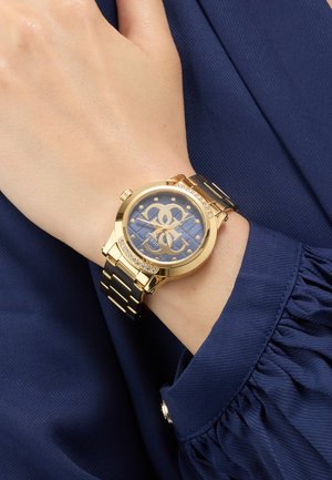 Orologio da polso in metallo oro e nero con quadrante blu decorato e cristalli, indossato su un polso sopra una camicia a maniche lunghe blu scuro.