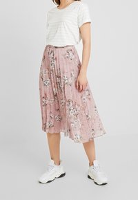 Kobieta w białej koszulce w paski, różowej, kwiecistej, plisowanej spódnicy midi i białych, masywnych trampkach, stojąca na jednolitym tle.