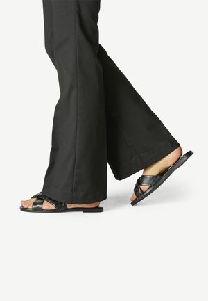 Sandales noires avec des brides texturées et une semelle plate, portées avec un pantalon noir à jambes larges. Les sandales présentent un design minimaliste et des brides réglables.