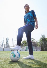 Sportska odjeća s plavim grafičkim dizajnom na tamnoj majici u kombinaciji s mornarsko plavim hlačama, naglašena ljubičastim prugama i bijelim nogometnim kopačkama.