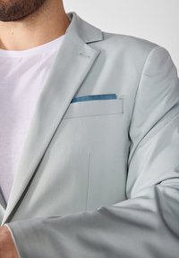Sacou gri deschis cu o textură netedă, cu reveruri și un batistă albastră în buzunarul de la piept. Purtați-l peste un tricou alb.