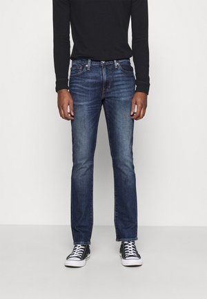 Man draagt donkerblauwe slim-fit jeans, een zwart shirt met lange mouwen en zwart-witte sneakers, staand tegen een effen witte achtergrond.