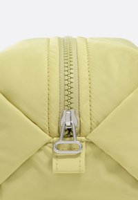 Żółta torba z pikowanej tkaniny z centralnym zamkiem błyskawicznym w srebrnym kolorze i teksturowym wykończeniem. Podkreślona geometrycznym przeszyciem.