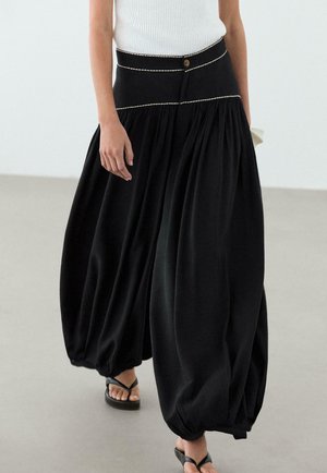 Donna che indossa una gonna balloon nera a vita alta con cuciture bianche, abbinata a una canotta bianca a coste senza maniche e sandali neri.